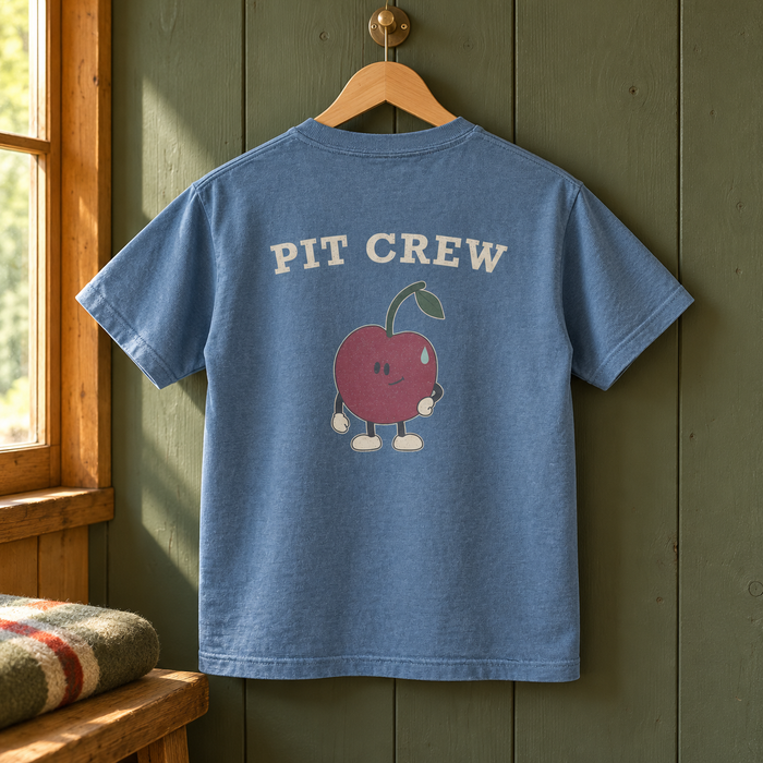 Camp Crew Youth Tee — Blue Jean — large back graphic on a wooden hanger in a warm camp cabin